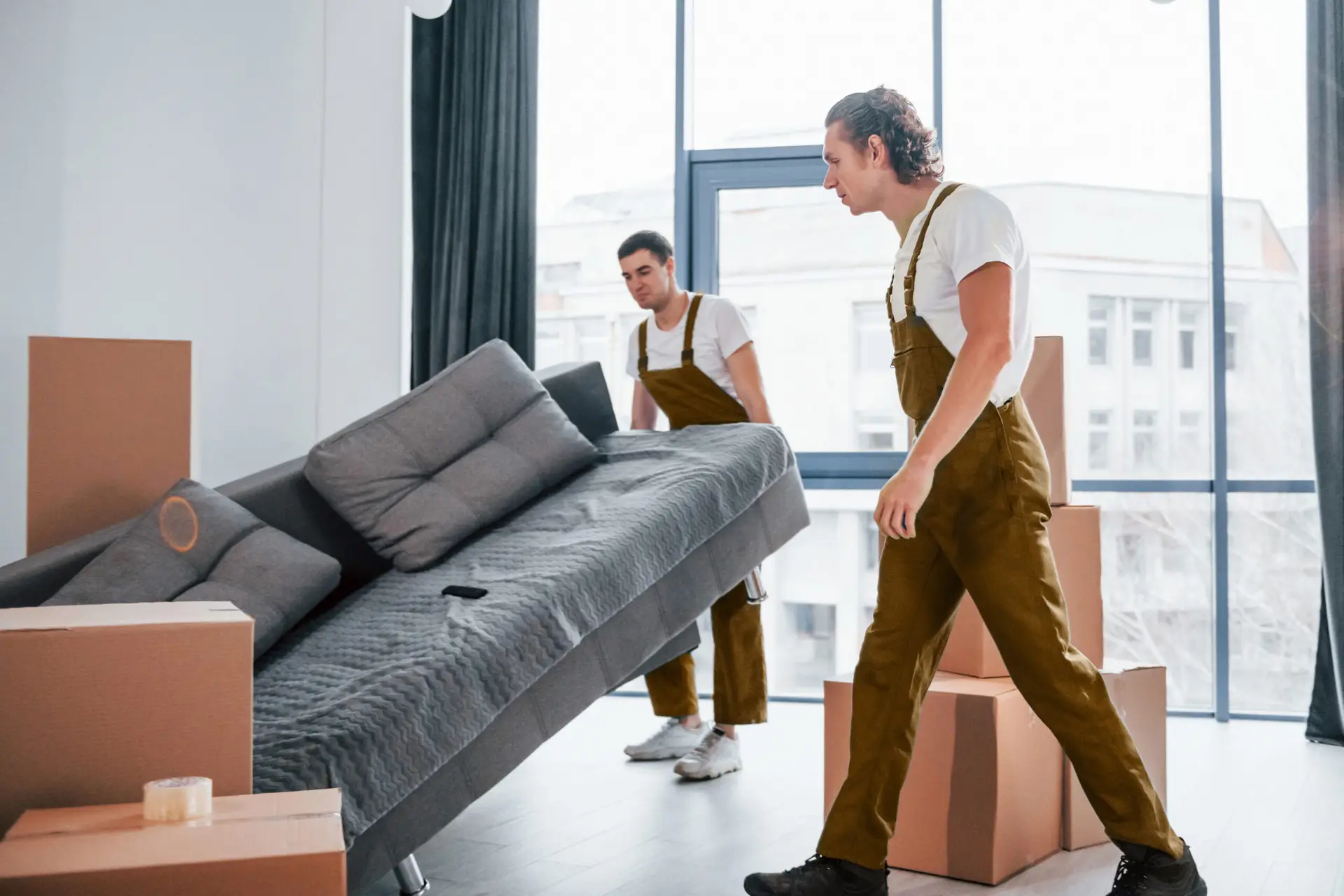 Two movers in brown overalls carrying a gray sofa in a bright, modern room with cardboard boxes, illustrating professional cleanout services in Vienna.